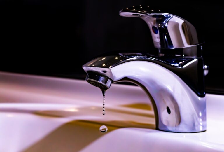 A person pouring tap water from a kitchen faucet into a clear glass pitcher on a countertop.
